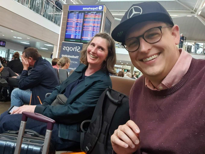 Dan and Hilary selfie in the airport. dan, glasses and black hat. hilary short dark hair, both smiling.