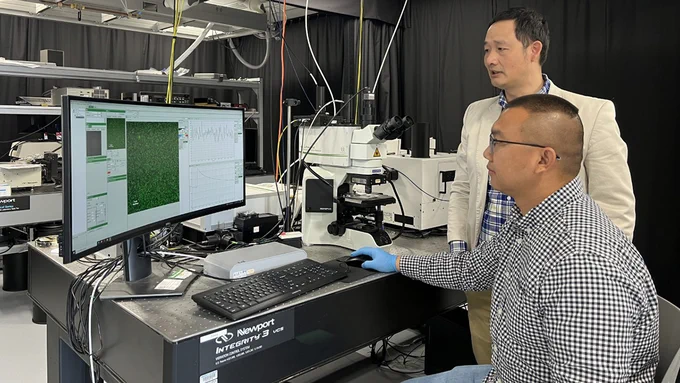Photo of Jinsong Huang (an East Asian man in a white blazer) and Ying Zhou (an East Asian man in a checked button-up shirt wearing lab gloves) sitting at a computer station in a lab