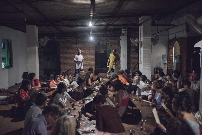 A photograph of lots of people sat around long wooden tables in a dimly lit room. They are listening to two people, who are standing up in the centre of the room, talking. 

Image credit: CLIMAVORE ON MANGROVES AND MUDFLATS BY COOKING SECTIONS AT COLOMBOSCOPE, PHOTOGRPAHY BY RUVIN DE SILVA.