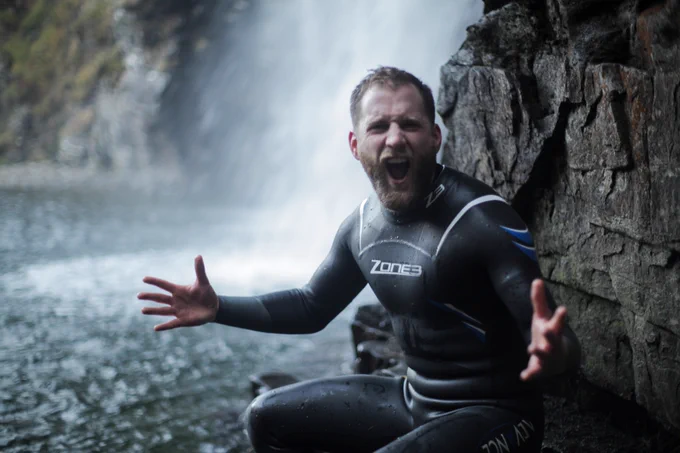 Calum Maclean sitting next to waterfall with arms wide, joyous face at River Douchary