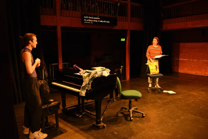 Two actors stand on a stage with a grand piano and some office chairs on it. A captioning unit can be seen hanging off the back wall. 