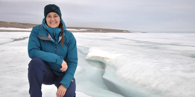 A smiling person with long brown hair tied back in a braid and wearing a blue snow jacket and pants and a blue winter hat kneels on a white ice field
