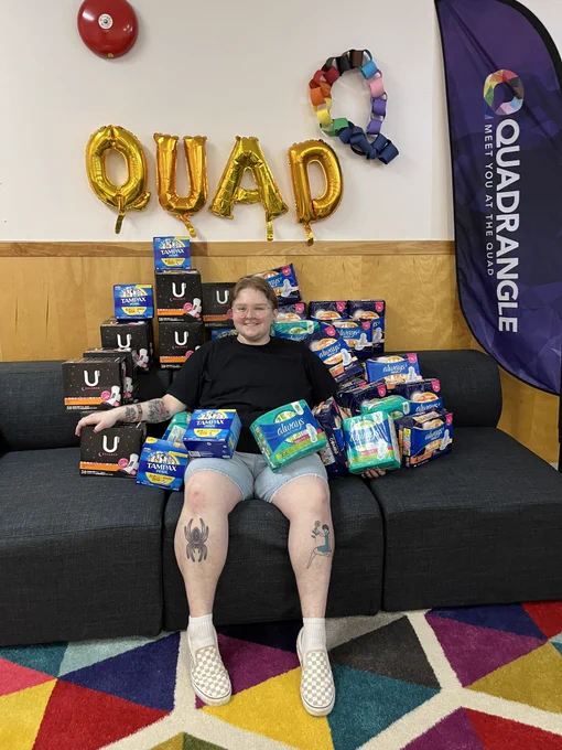 Holland sitting on a couch with period products. There is a balloon that reads "Quad" on the wall. 