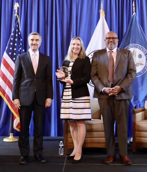 Assistant Secretary Venkataraman and two others pose standing for a photo. In the middle, a woman holds an oval-shaped award. In the background are the U.S., Department of State and Department of Commerce flags.