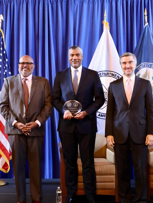 Assistant Secretary Venkataraman, Deputy Assistant Secretary Ian Saunders, and Assistant Secretary Toului pose for a photo standing. Deputy Assistant Secretary Saunders, standing in the middle, holds an oval shaped award. In the background are the U.S., Department of State and Department of Commerce flags.