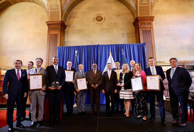 A group photo featuring several Department of Commerce and Department of State officials, including Assistant Secretary Venkataraman, Assistant Secretary Toului, Deputy Assistant Secretary Saunders and Deputy Assistant Secretary De Falco. All are standing and some are holding framed award certificates.