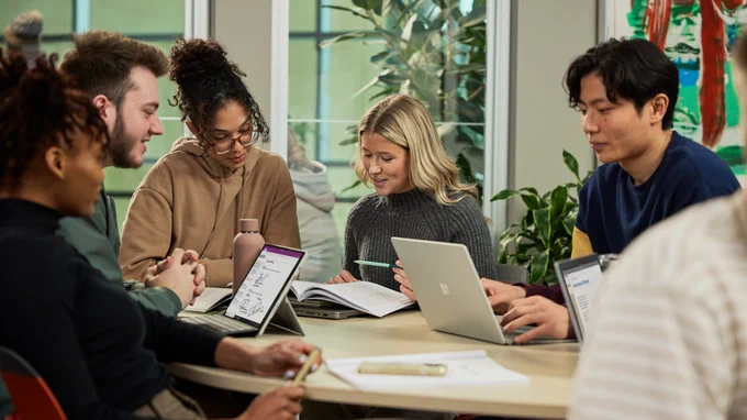 A group of young people at their laptops sit together at a round table talking with each other.