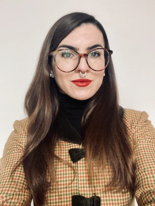 front facing profile photo of a young female-presenting professional with long brown hair, white skin, round glasses, red lips, and a bull ring nose ring in a checkered jacket on a white background
