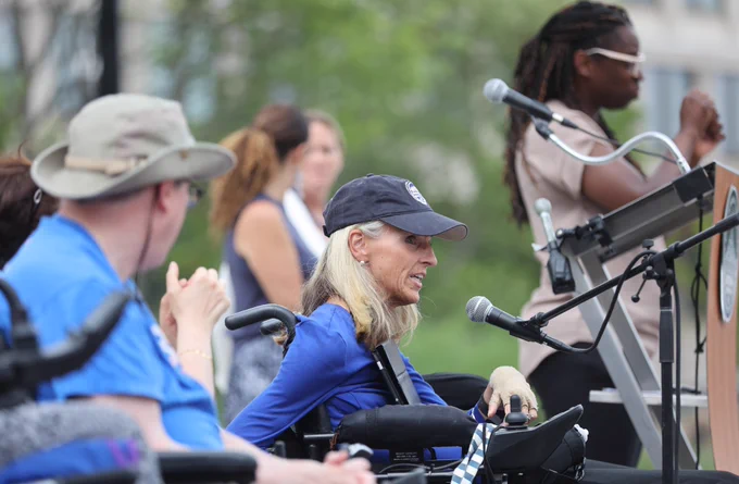 Commissioner of the Boston Disabilities Commission Kristen McCosh, speaks behind a podium. Using a black wheelchair, with a black microphone positioned in front of her, Commissioner McCosh looks forward as she speaks, wearing a blue shirt and a black baseball cap decorated with the City of Boston logo in front in white. To her left sits a community member using a wheelchair, wearing a grey hat, dark glasses, and a light blue shirt, who is watching the Commissioner as she speaks. To the right of the commissioner stands an ASL interpretor, wearing a beige short sleeved shirt and white glasses, their hands gesturing in front of them.