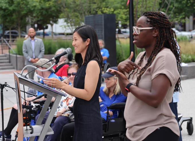 Mayor Wu stands at a podium, smiling at the audience, wearing a navy blue, sleeveless dress. Beside her, in the foreground, is an ASL interpretor, their right index finger pointing at their chest, their left hand, palm open, under their right hand. The interpretor wears white sunglasses and a beige colored t-shirt. Behind them sits the Commissioner of the Boston Disabilities Commission, Kristen McCosh, who uses a wheelchair, with her arms crossed in front of her, wearing a dark baseball cap.
