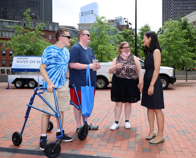 Mayor Wu speaks with three other community members, standing on the bricks of City Hall Plaza. From right to left, stand: the Mayor, wearing light colored shoes and a dark sleeveless dress; a community member wearing dark rimmed glasses, a pink mask at their chin, a sleeveless black and pink shirt, a black knee-length skirt, and white shoes with dark socks; a community member wearing dark glasses, navy blue shirt with red pants, and holding a light blue bag in their right hand while also carrying a cup; and a community member wearing dark glasses, a tie-dyed bright blue shirt, khaki pants, black socks, with one hand in their pocket while the other grips a blue walking aid.