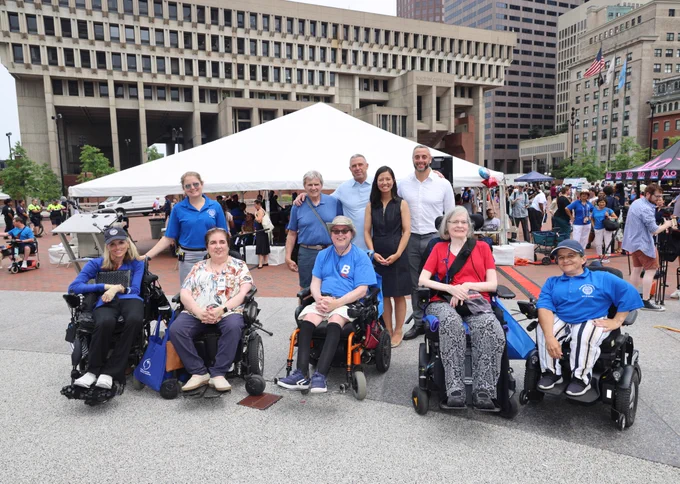 A group of 10 people — including Mayor Wu, Boston’s Chief of Streets Jascha Franklin-Hodges, Boston’s Disabilities Commission Commissioner Kristen McCosh, and other City Hall workers — are pictured in front of a white tent placed on City Hall Plaza, with 5 persons in wheelchairs and the rest standing behind them. Boston City Hall, a beige windowed building with brutalist architecture, is in the background. Towards the right of the photo, to the right of the white tent behind the group, stands a crowd of people, walking along City Hall Plaza.