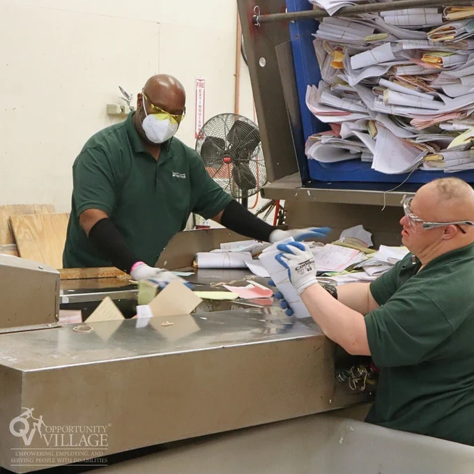 Two people standing on either side of a large paper shredding conveyer belt. There is a large bucket of documents to their side ready to be emptied into the shredder. 