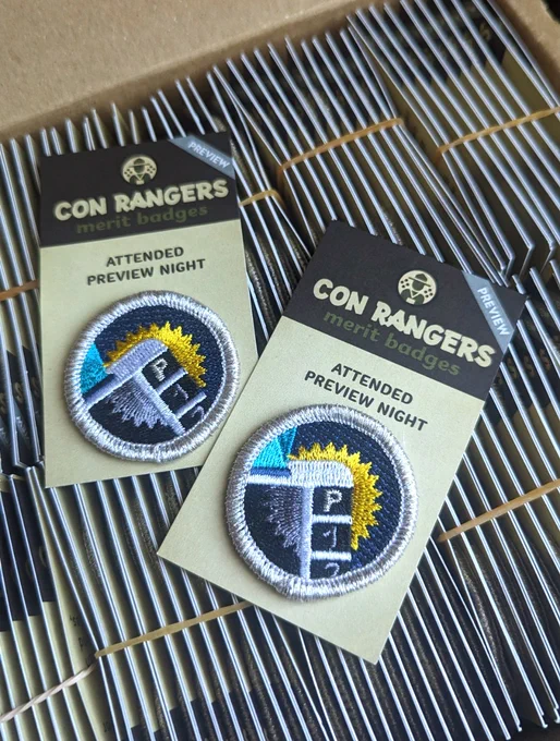 A Con Rangers badge for "Attended Preview Night" - silver metallic thread around a con badge that has a starburst behind the P for Preview Night, on top of a box of lots of badges