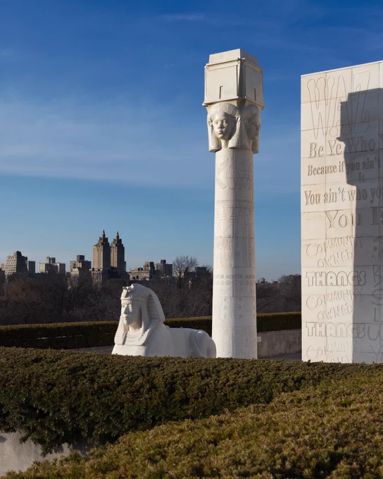 A photograph depicts The Met Rooftop Commission against a clear, blue sky. The commission is an Egyptian inspired structure which includes a sphinx-like figure, a large column with a portrait carved in relief at the top, and the corner of a building which is inscribed with graffiti. 