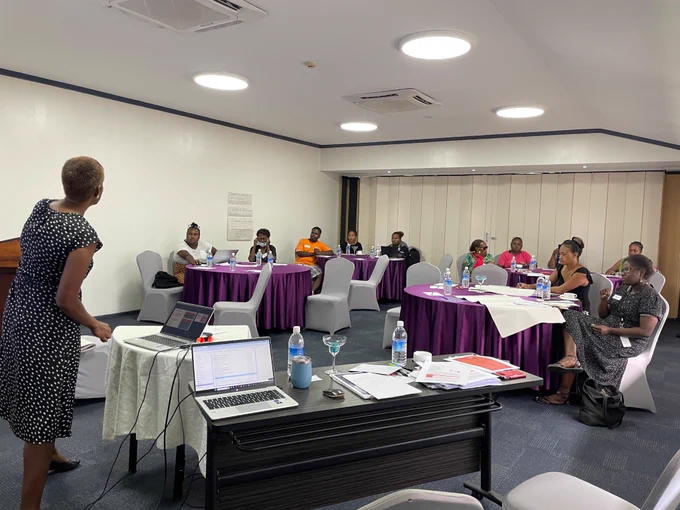 A group of people are sitting in groups at round tables listening to a presentation. A woman is at the front of the room presenting to the group. Many of the people in the room are Solomon Islanders.  