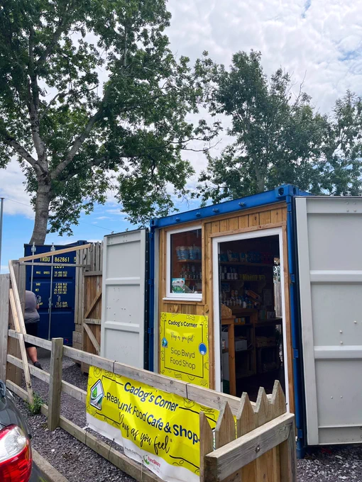 A few shipping containers converted into shops under trees. Big yellow banners read “Cadog’s Corner Cafe and Shop”