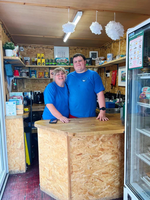 Two smiling volunteers in blue Cadog’s Corner T-shirts. Sue on the left is a lady with a fringe and Josh on the right is a head taller with dark hair. They stand behind the counter in the container shop.