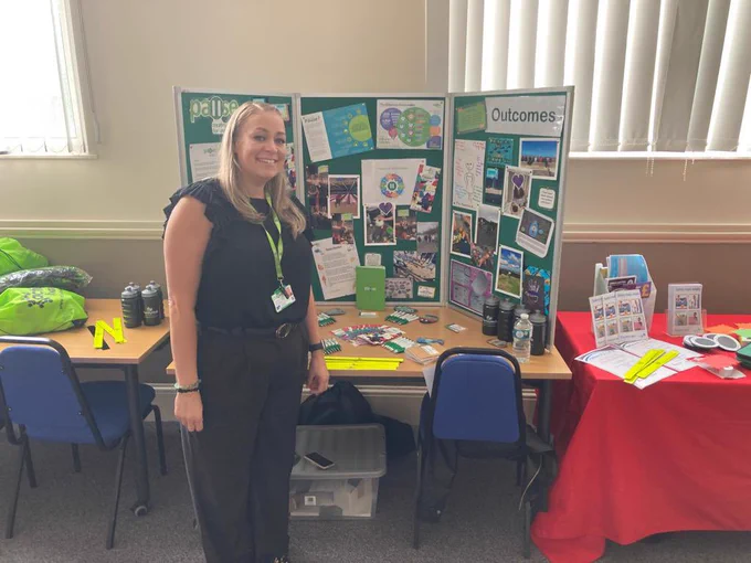 Practice Lead, Lindsey Clarke, standing in front of a table. Lindsey is a white woman with blonde hair. She is wearing a black top and trousers. On the table behind her is a board with information about Pause.