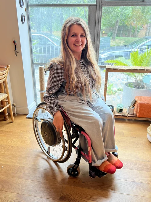 katie, a white woman with blondish hair, sitting in her pink manual wheelchair in the exact same pose as the barbie in the previous photo.