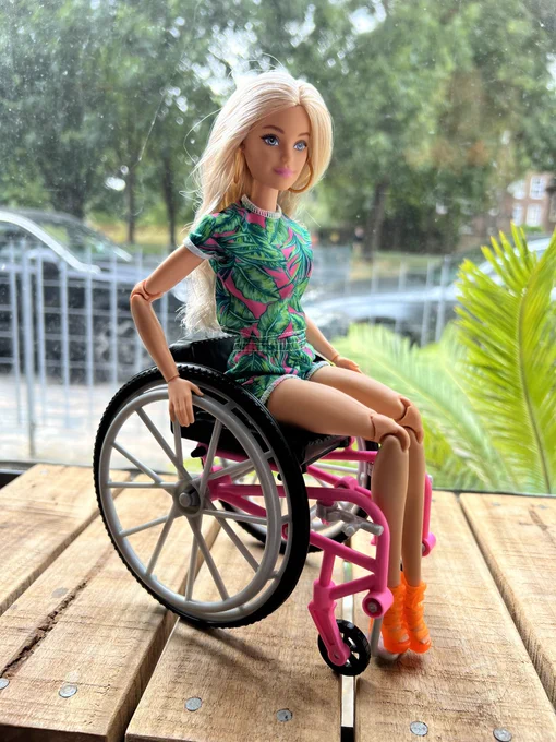 a barbie doll with light skin and blonde hair sitting in her barbie wheelchair which is a pink manual wheelchair.