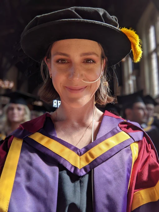 Photo of a white woman with an NJ tube smiling wearing a PhD graduation gown and Tudor bonnet. 