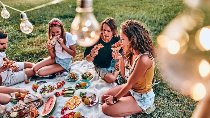 Group of young adults sitting around a picnic eating pizza and fruit.