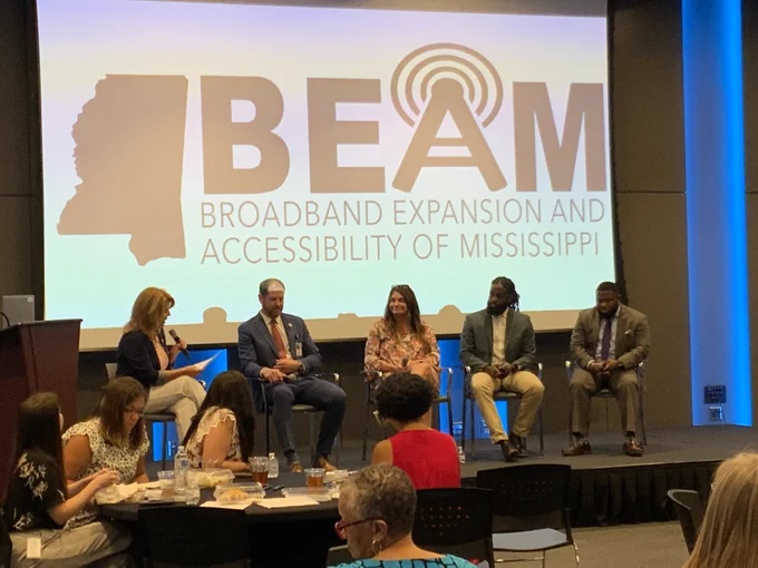A group of people sit on a stage before a display showing the logo of BEAM: Broadband Expansion and Accessibility of Mississippi.