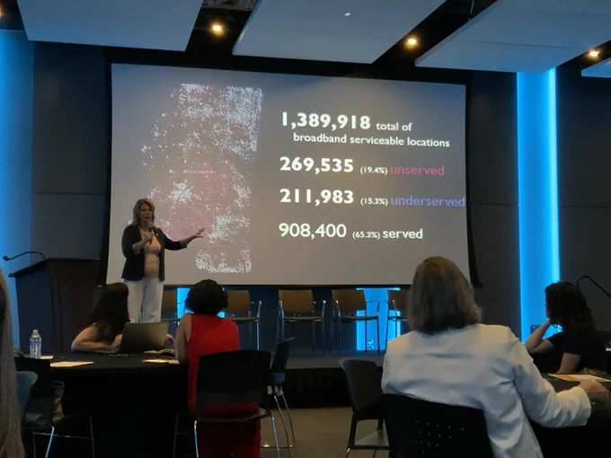 A woman stands in front of a presentation. The slide reads: 1,3,89,918 total of broadband serviceable locations. 269,535 (19.4%) unserved. 211, 983 (15.3%) underserved. 908,400 (65.3%) served.