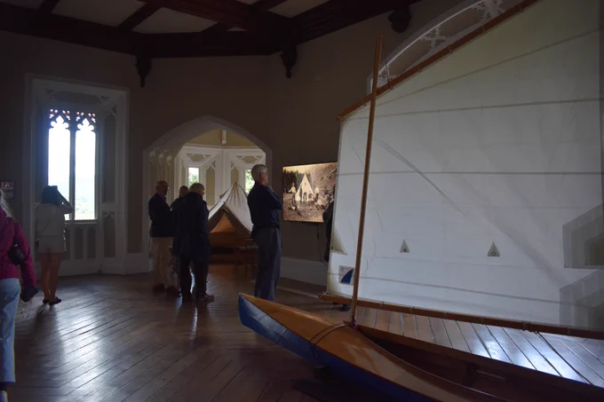 A wide shot of the Brought to Light exhibition, with visitors milling around. In the centre of the large room is a bright blue sailing boat with a white sail