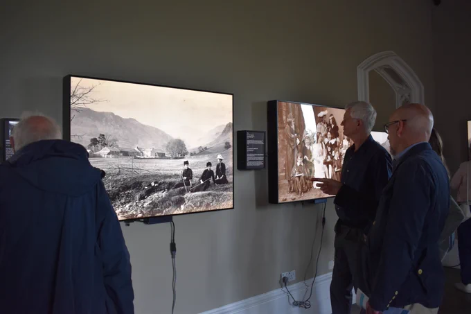 Harvey, the curator at Wray Castle, shows The Armitt's team photos from the Herbert Bell section of the Brought to Light exhibition. Harvey, a bald, bearded man in a blue jacket and glasses, stands with Maurice George on the left and Peter Lansberry on the right in front of a lightbox. In the light box is a black and white image of three female walkers from the nineteenth century resting on a river bank