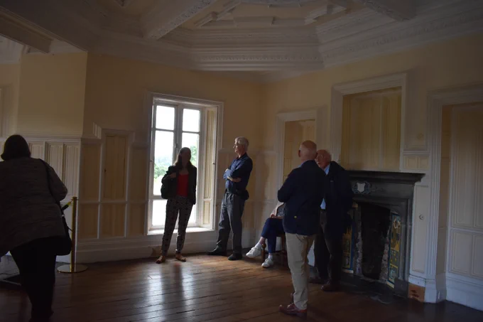 The Armitt team with Harvey in another room in Wray Castle. The room is white and cream in colour, with ornate moulding and a fireplace on the right hand side. Next to the fireplace stands Harvey and Tony Lonton, with volunteer Judy sat on a chair to the left, her face obscured. Trustee Peter Lansberry stands to the left of the window, looking out. Armitt manager and curator Faye Morrissey stands in front of the window, with admin assistant Sue on the far left