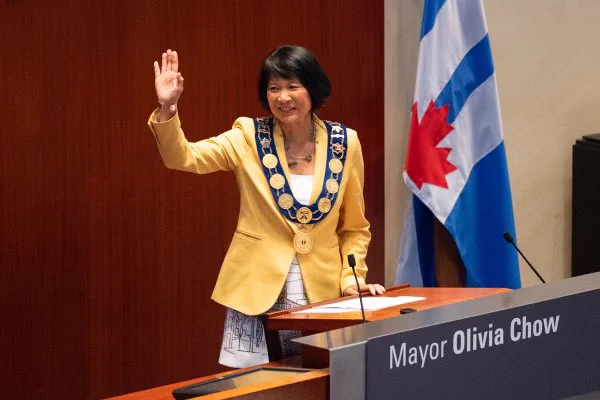 Mayor Olivia Chow stands at the speaker’s dias in the Council chamber and waves to people in the gallery.