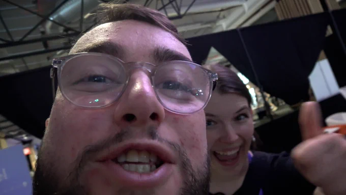 A close up of Mini Muka's face. He's mid-sentence, while Sarah smiles and holds a thumbs up from behind him.