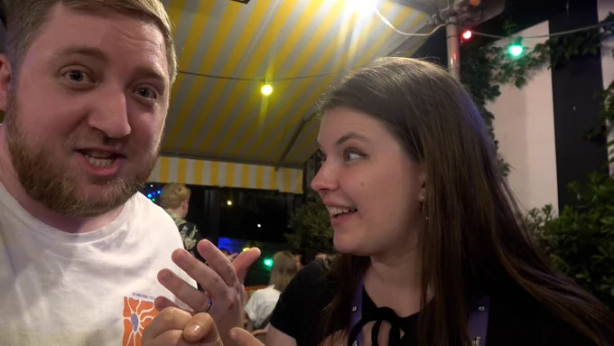 Sarah holds an invisible microphone up to Martyn's face. He's speaking intently, she looks amazed. They're inside a tent with a yellow and white striped roof.
