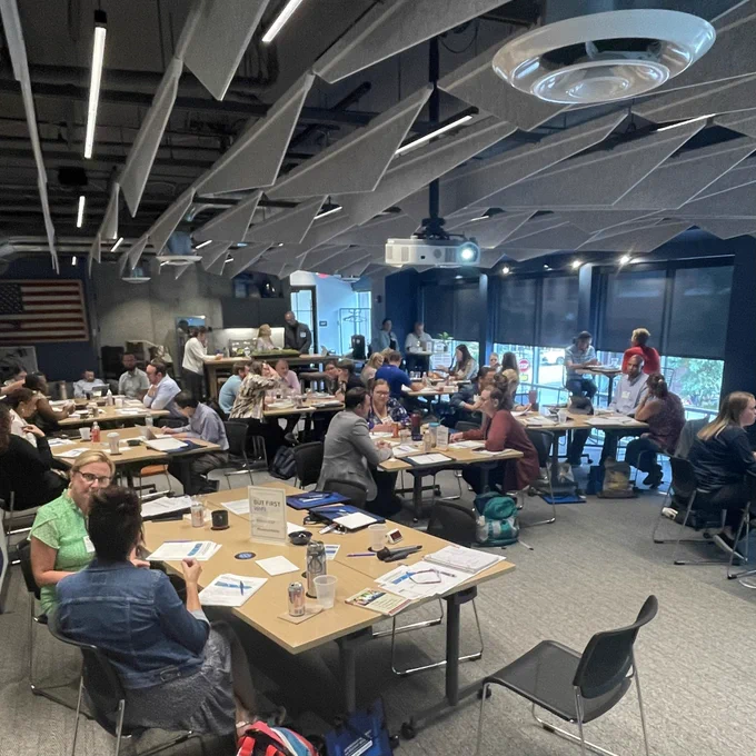 Small groups meeting at tables in a collaborative work space. Notebooks and worksheets at each table. Approximately 40 people in groups of 2-5.