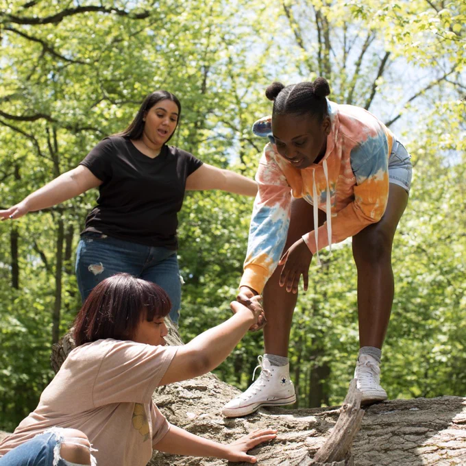 Three youth of color thriving outdoors. In the foreground, a Black youth helps another youth of color over a large log. A third Latinx youth walks on the log, arms outstretched to balance.