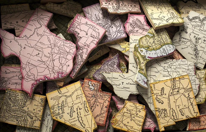 Jumbled pile of USA state-shaped puzzle pieces. Each piece is a solid in color with black text displaying the state name and major roadways, as if they were cut from a roadmap. The pieces are displayed in a range of muted colors including light pink, yellow, green, and orange. The State of Michigan piece also includes the surrounding water in blue. 