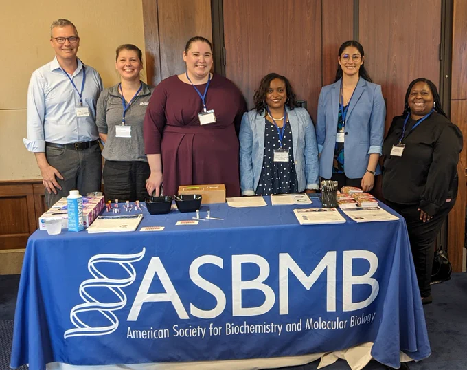 Six ASBMB committee members and staff members stand behind a table with a blue table cloth that reads ASBMB at the Congressional Life Sciences Fair at the Rayburn House building in Washington, D.C. 