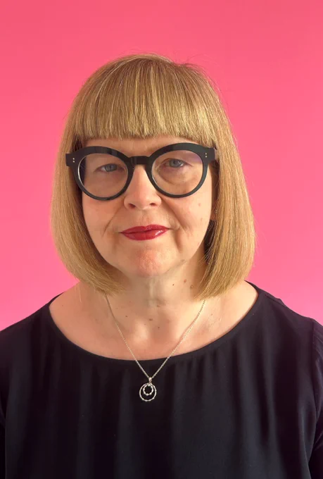 Photograph of Annabel Turpin, a white woman in her early 50s, with a blond bob cut. She is wearing black glasses, red lipstick, and a dark purple top, and is standing in front of a pink background.