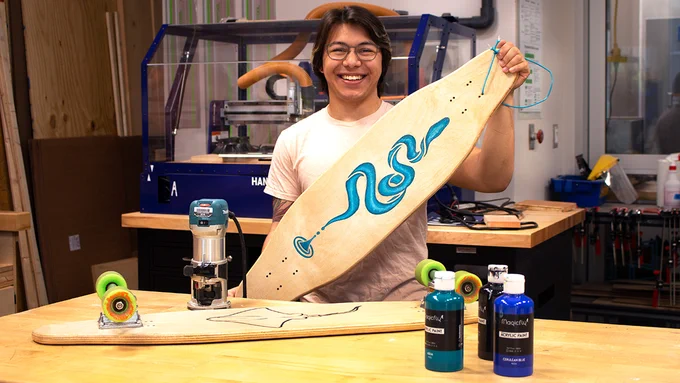 Levi Tox holds his longboard, which features a snake design, in the Murray makerspace. Another skateboard is on the table in front of him.