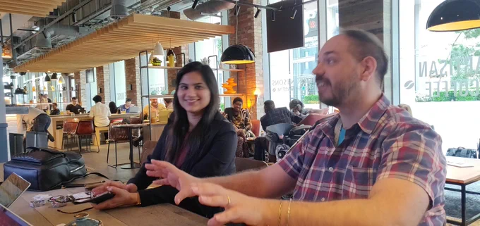 Location: Coffee shop. a woman smiling on the left and a man on the right with his hands slightly raised as he's moving them whilst talking.