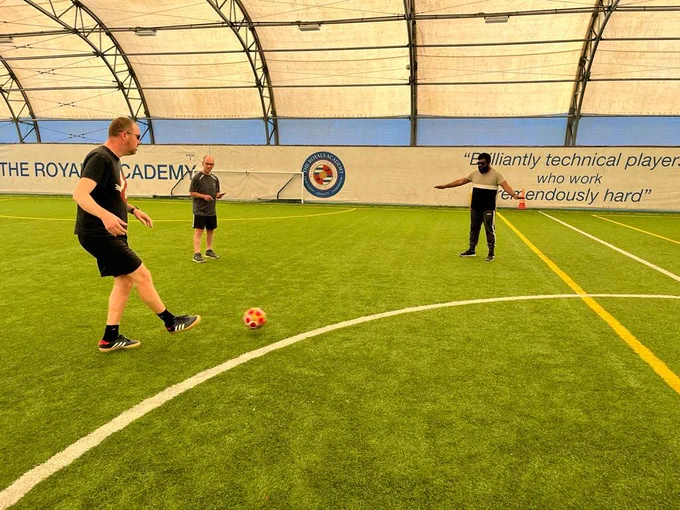 3 people are on the astroturf in the Dome at Reading Football Club. They are all wearing dark glasses and one person is kicking the ball tp another person. 