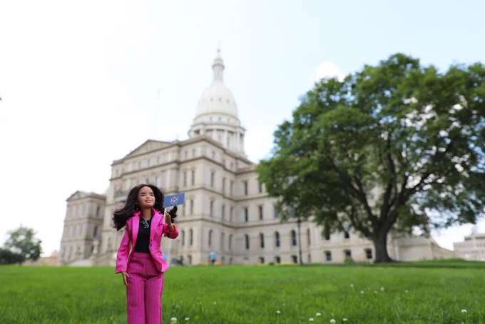 A Barbie version of Michigan Governor Gretchen Whitmer stands in front of the Michigan State Capitol holding a Michigan flag
