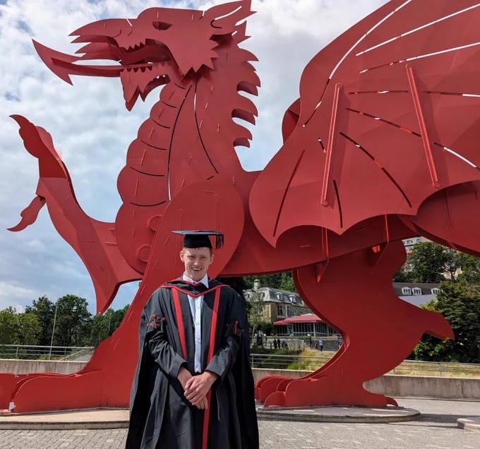 A man wearing graduation cap and gown, stood in front of a red dragon statue