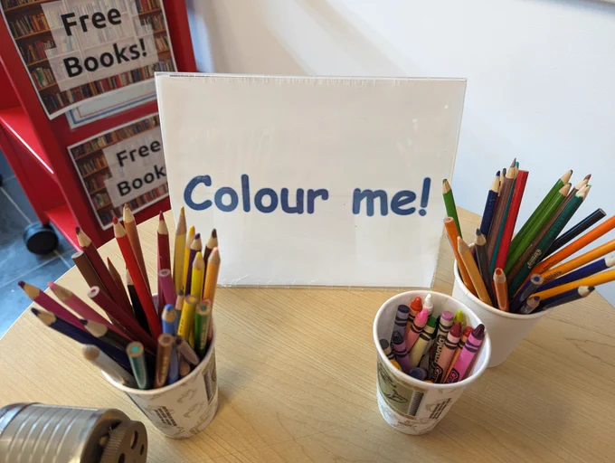 A cup of crayons and two cups of pencil crayons on a table just inside the Charles Hall Library doors, with a sign that says "Colour me!"