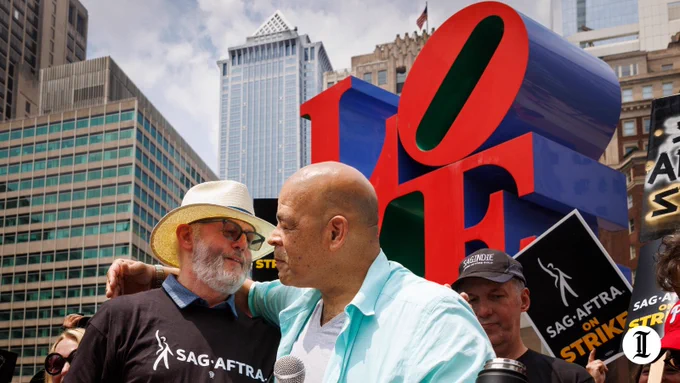 Actors David Morse (left) and Brian Anthony Wilson (right) embrace at the podium while the LOVE sign looms in the background.