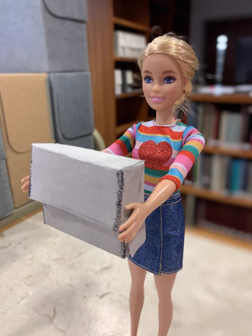 Barbie doll wearing a striped sweater with a heart and a jean skirt stands holding a fibredex box.
