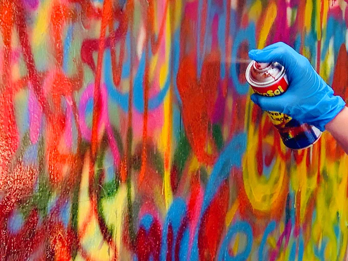 Eine Hand in einem blauen Handschuh sprüht Graffiti auf eine bereits bunte Wand.