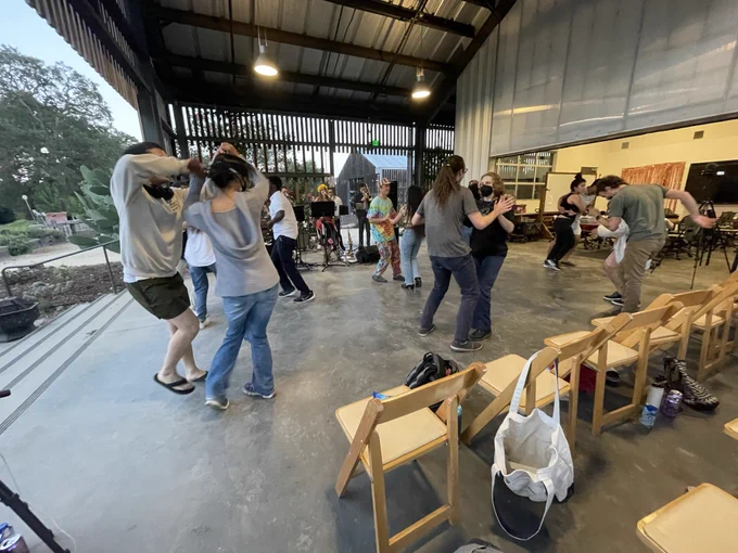 several people dancing in a modern farm. it is a beautiful sunset. some folks are masked. the singer is wearing sunflower-type garland in her hair.